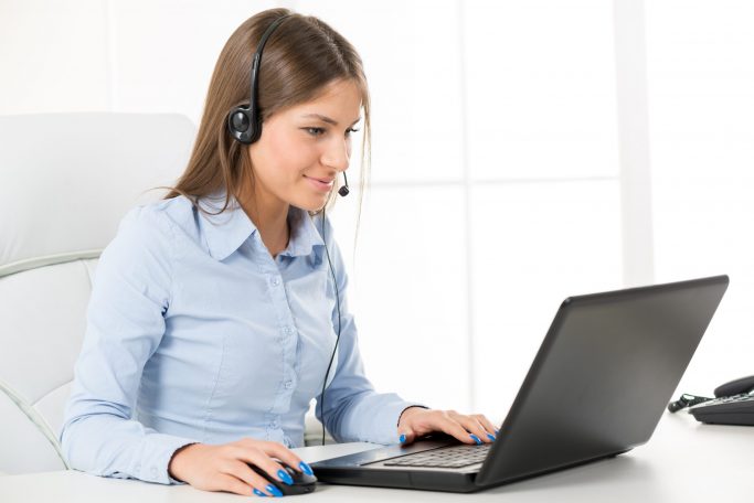 Young,Businesswoman,With,Headset,,Smiling,,Typing,On,A,Laptop,In